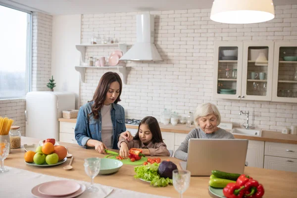 Mom with daughter making salad near grandmother with laptop. — 스톡 사진