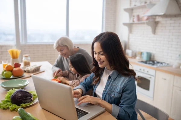 Moeder op laptop in de keuken, grootmoeder met kleindochter snijden salade. — Stockfoto