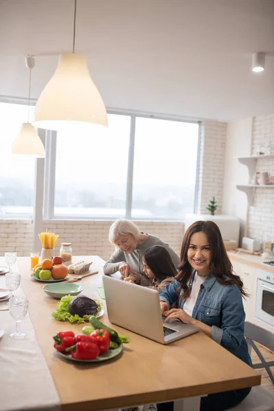 Mom with laptop, grandmother and granddaughter in morning in kitchen. — Stock Photo, Image
