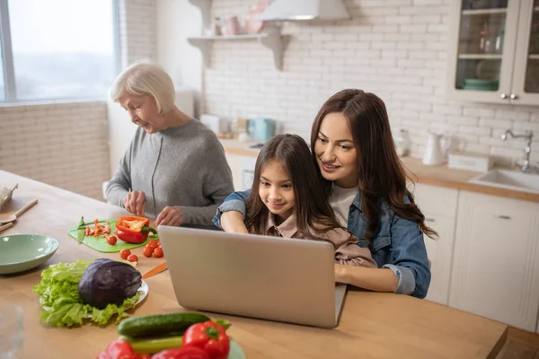 Mor med datter på laptop, bedstemor tilbereder mad i køkkenet . - Stock-foto