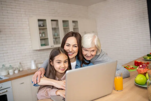 Nagymama, anya, lánya egy laptopnál ölelkeztek a konyhában.. — Stock Fotó