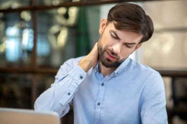 Bearded man in a blue shirt having pain in his neck