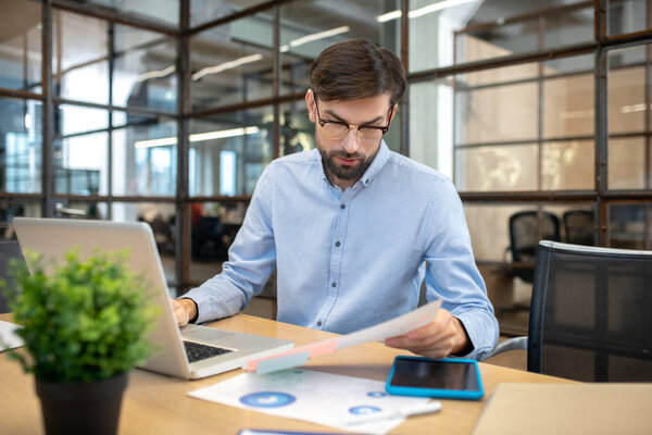 Bearded man in a blue shirt wearing eyeglasses working in the office and looking involved