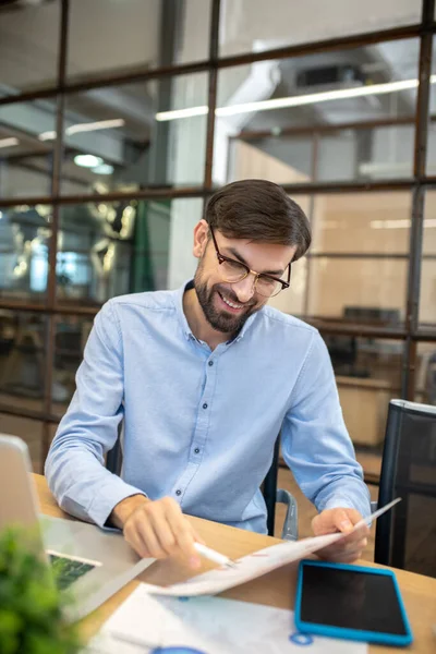 Szakállas férfi kék ingben szemüvegben, az irodában dolgozik papírokkal. — Stock Fotó