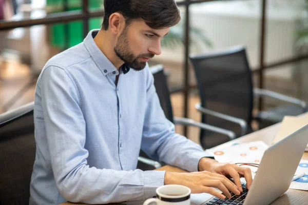 Jeune homme barbu dans une chemise bleue regardant concentré tout en travaillant sur un ordinateur portable — Photo