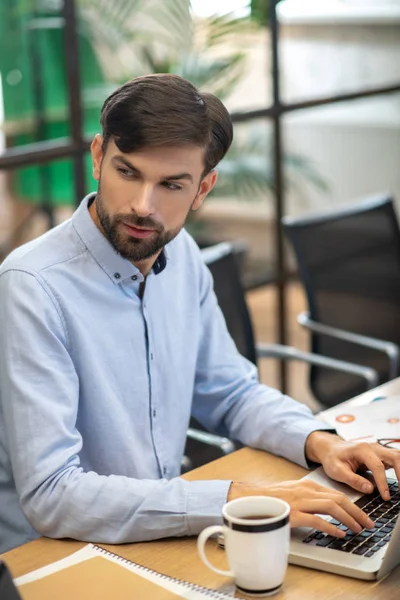 Fiatal szakállas férfi kék ingben, komolynak tűnik, miközben egy laptopon dolgozik. — Stock Fotó