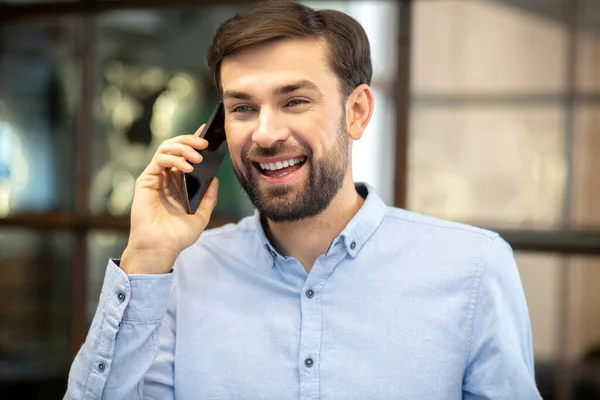 Молодой бородатый мужчина в синей рубашке разговаривает по телефону и чувствует себя потрясающе — стоковое фото