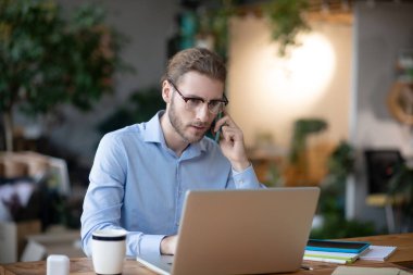 Bilgisayarda çalışan genç bir iş adamı akıllı telefondan konuşuyor..