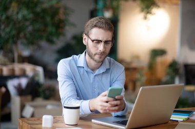 Elinde akıllı telefon olan genç bir adam bilgisayarın başında oturuyor..