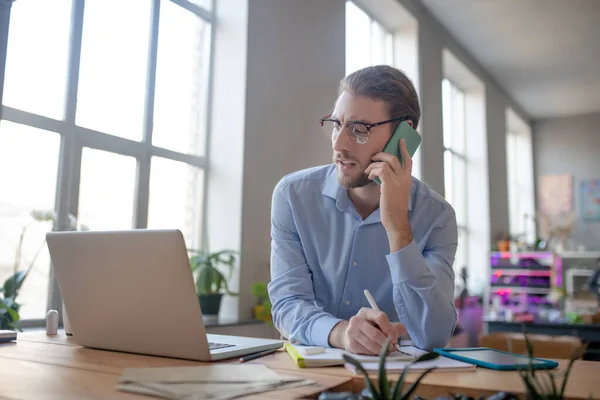 Joven reflexivo hablando en el teléfono inteligente, tomando nota en el proyecto . — Foto de Stock