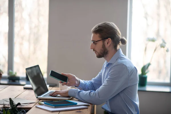 Kontorsarbetare sitter på laptop med smartphone i handen. — Stockfoto