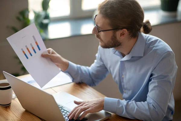 Ernster Mann, der am Laptop arbeitet und auf das Diagramm in seiner Hand blickt. — Stockfoto