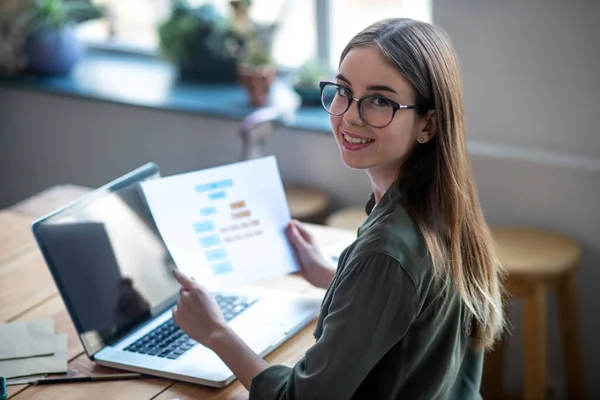 Flicka i glasögon framför laptop med diagram i händerna. — Stockfoto