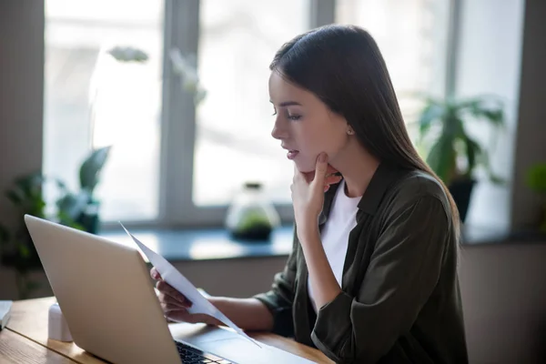 Flicka sitter framför laptop noggrant tittar på pappersdokument. — Stockfoto