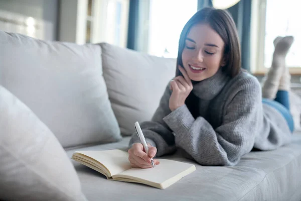 Junges Mädchen im grauen Pullover, das Notizen macht und glücklich aussieht — Stockfoto