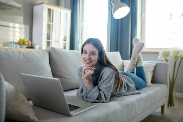 Young girl in a grey sweater laying on the sofa and chatting online — Stock Photo, Image