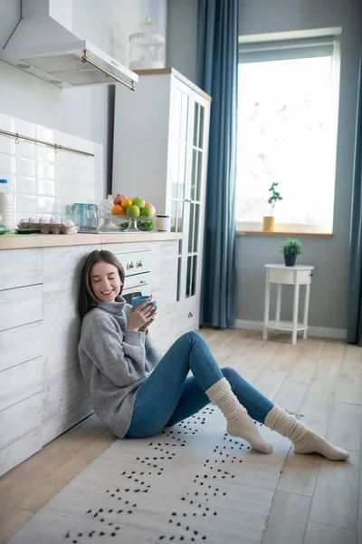 Dark-haired girl in a grey sweater sitting on the floor and having coffee — Stok fotoğraf