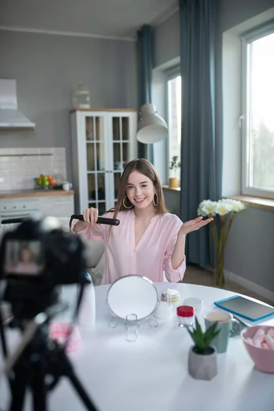 Sonriente chica bonita con una camisa rosa sosteniendo un rizador en sus manos — Foto de Stock