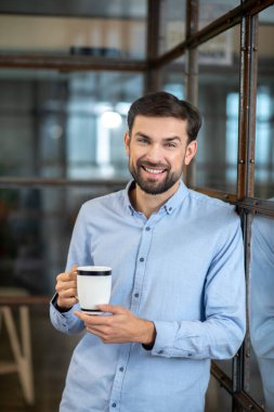 Young bearded man in a blue shirt smiling and having coffee