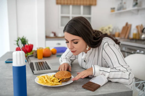 Tmavovlasá žena v pruhované blůze, která jí hamburger a bramborové lupínky — Stock fotografie