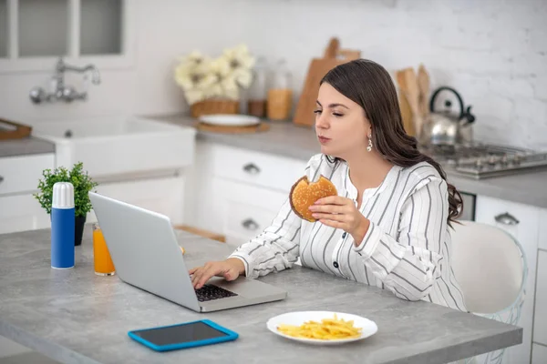 Σκούρα μαλλιά γυναίκα σε μια ριγέ μπλούζα τρώει ένα burger και εργάζεται — Φωτογραφία Αρχείου