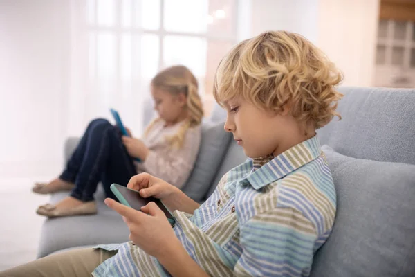 Niño y niña sentados en el sofá y jugando con gadgets —  Fotos de Stock