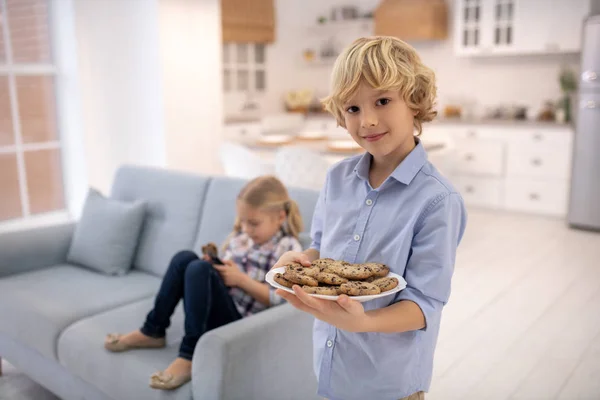 Boy standing with plate of biscuits, girl sitting on sofa — Stock Photo, Image