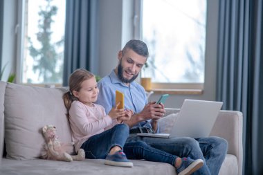 Daughter showing smartphone screen to dad, with smartphone and laptop.