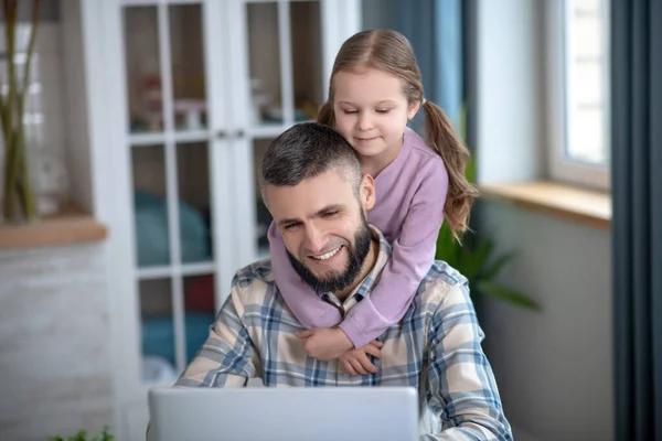 Dad working with laptop, little daughter hugs him from behind.
