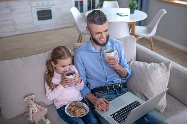 Dad and daughter looking at a laptop drinking tea eating cookies. — 스톡 사진