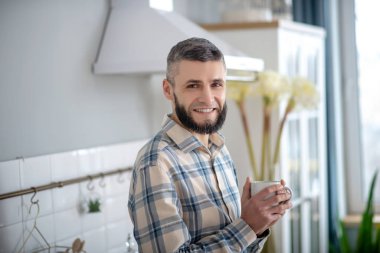 Smiling young bearded man standing with a white cup.