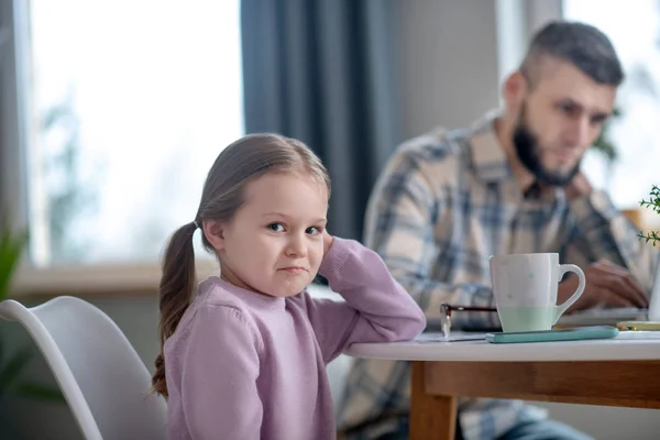 Klein meisje zit aan tafel droevig glimlachend, papa bezig. — Stockfoto