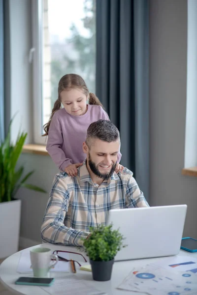 Dad sitting working at laptop, little daughter behind daddys back. — Stock Photo, Image