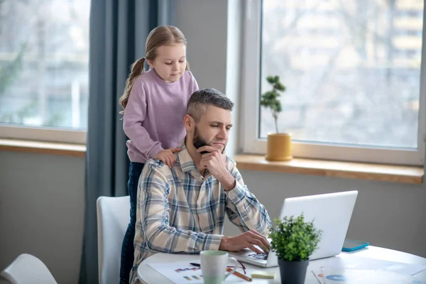 Sentado focado pai, filha de pé atrás, eles estão olhando para laptop . — Fotografia de Stock