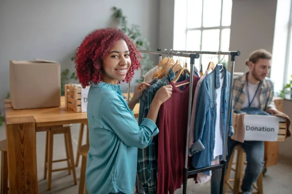 Chica joven cerca de rack con ropa hombre con caja de donación . — Foto de Stock