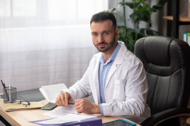 Ofisindeki masada oturan beyaz cüppeli genç sakallı doktor.