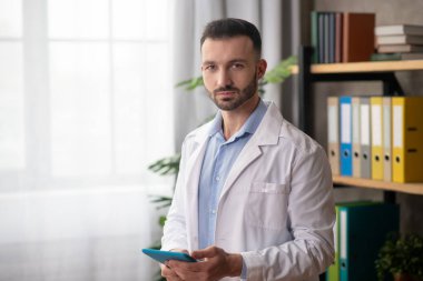 Beyaz cüppeli genç sakallı doktor elinde bir tablet tutuyor.