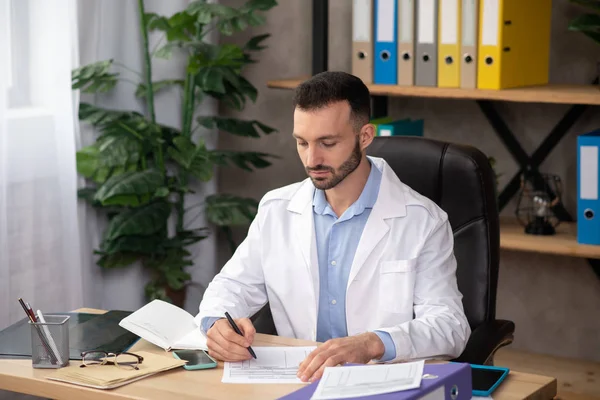 Jeune médecin barbu en robe blanche travaillant avec des papiers dans son bureau — Photo