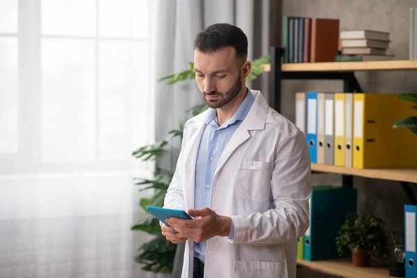 Joven médico barbudo con una bata blanca leyendo algo en una tableta — Foto de Stock