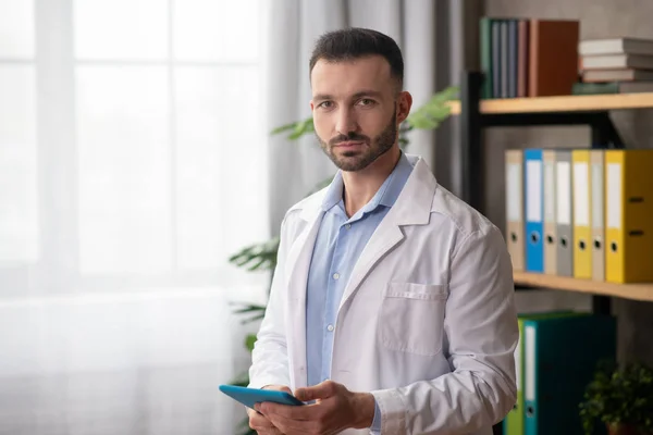 Joven médico barbudo con una bata blanca sosteniendo una tableta en sus manos — Foto de Stock