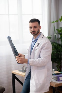 Beyaz cüppeli genç sakallı doktor MR sonuçlarını elinde tutuyor.