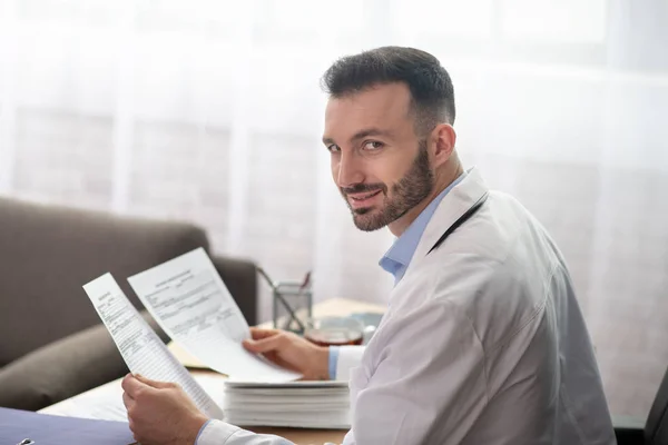 Donkerharige bebaarde arts in een wit gewaad ziet er vrolijk uit — Stockfoto