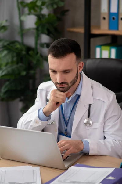 Mladý vousatý doktor vypadá vážně pracuje na notebooku — Stock fotografie