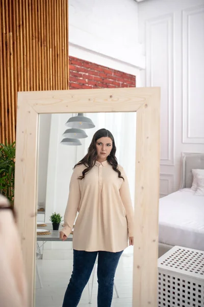 Young woman in a beige blouse looking at herself in the mirror — Stock Photo, Image