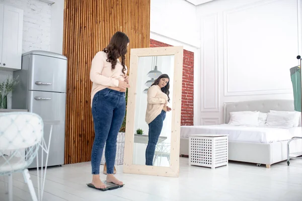 Молодая женщина в бежевой блузке, стоящая на весе и смотрящая в зеркало — стоковое фото
