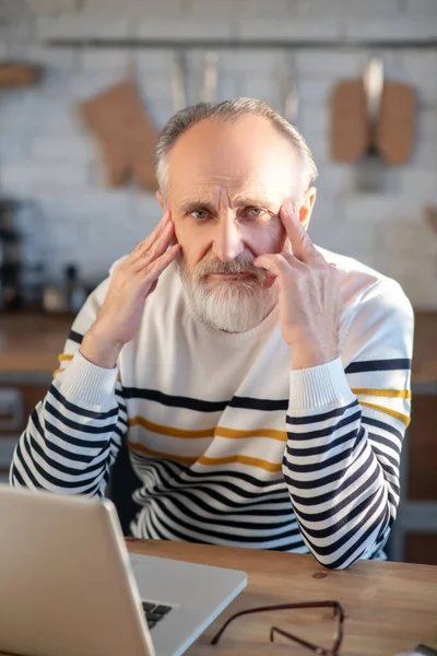 头发灰白、留着胡子、拿着笔记本电脑坐在桌旁工作的老年人 — 图库照片