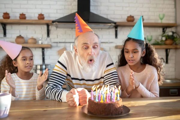 Homem barbudo com um chapéu de aniversário soprando as velas em seu bolo bday — Fotografia de Stock