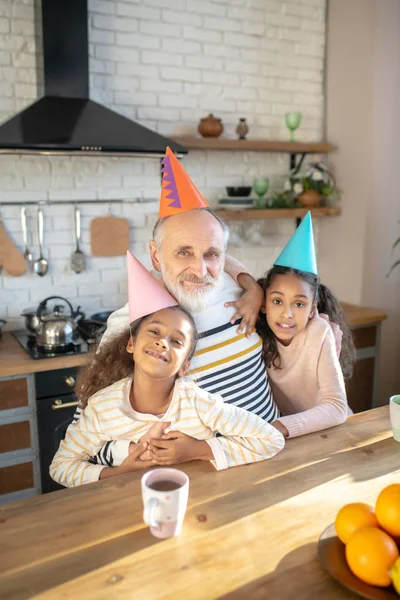 Två mörkhyade tjejer i fladdermössor som är nöjda med sin farfar. — Stockfoto