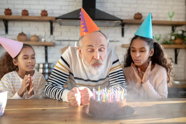 Bearded man in a birthday hat blowing down the candles on the cake