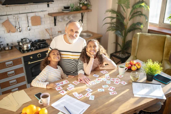 Två mörkhyade flickor leker ordlek med sin farfar och ser leende ut. — Stockfoto